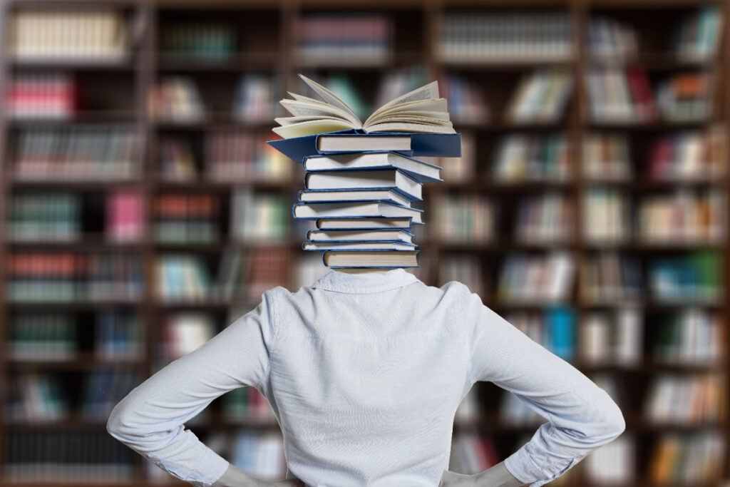 Corpo feminino de costas diante de uma prateleira de livros e no lugar da cabeça há uma pilha de livros.