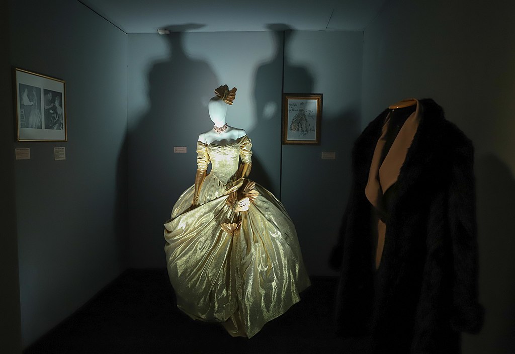 Vestido longo feminino do século XIX à esquerda e à direita batina de padre. A foto é em um ambiente escuro e com sombras projetadas na parede ao fundo.