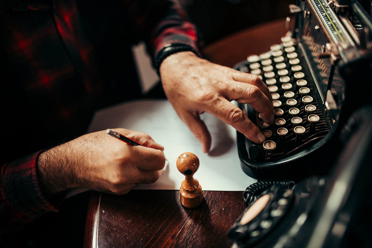 Pessoa com a mão esquera sobre teclas de uma máquina de escrever e mão direito escrevendo em um folha em branco sobre a mesa