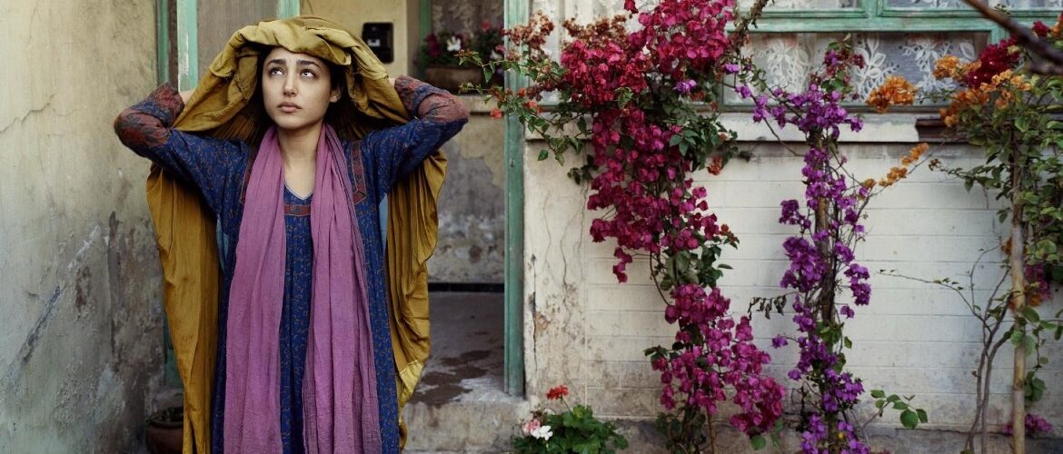 Mulher afegã com mão na cabela em frente a uma casa muito simples, com a porta aberta e uma pequena árvora com folhas rosas.
