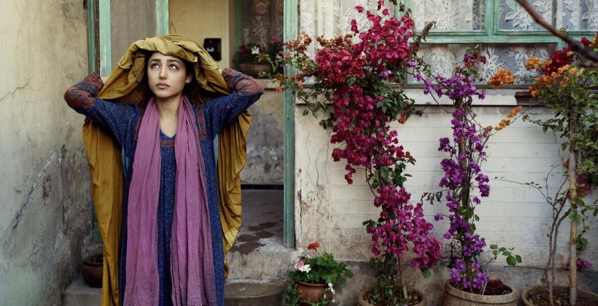 Mulher afegã com mão na cabela em frente a uma casa muito simples, com a porta aberta e uma pequena árvora com folhas rosas.