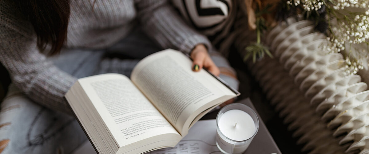 Pessoa segurando um livro no centro da imagem, com papeis e uma vela aromática sobre a mesa.