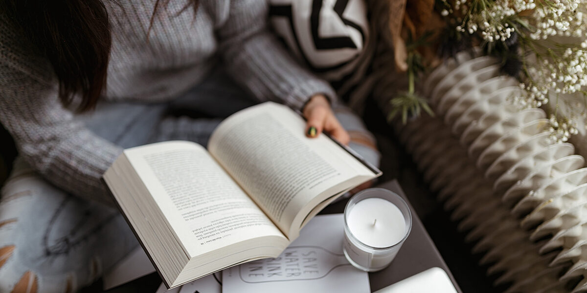 Pessoa segurando um livro no centro da imagem, com papeis e uma vela aromática sobre a mesa.