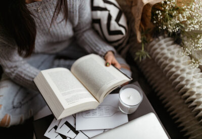 Pessoa segurando um livro no centro da imagem, com papeis e uma vela aromática sobre a mesa.