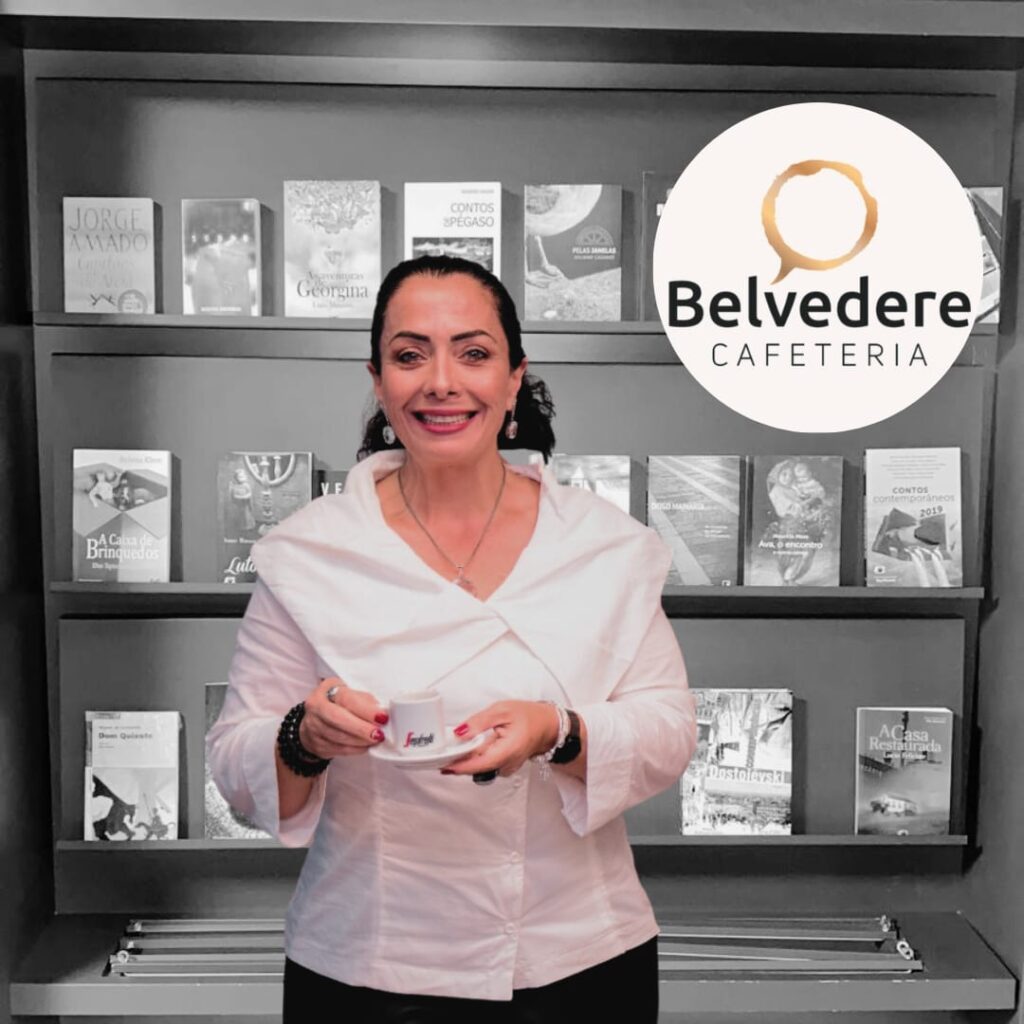 No primeiro plano Tatiana Cibele Mendonça Pereira proprietária do Belvedere Café Lounge, no Hospital Baía Sul, segurando uma xícara de café e, ao fundo, uma prateleiras de livros para empréstimo em preto e branco.