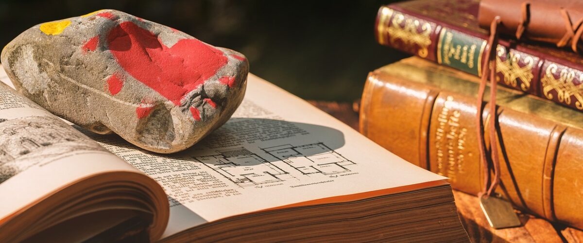 Livro aberto com uma pedra em cima pintada a superfície de vermelho. Ao lado três livros fechados, aparecendo a lombada.