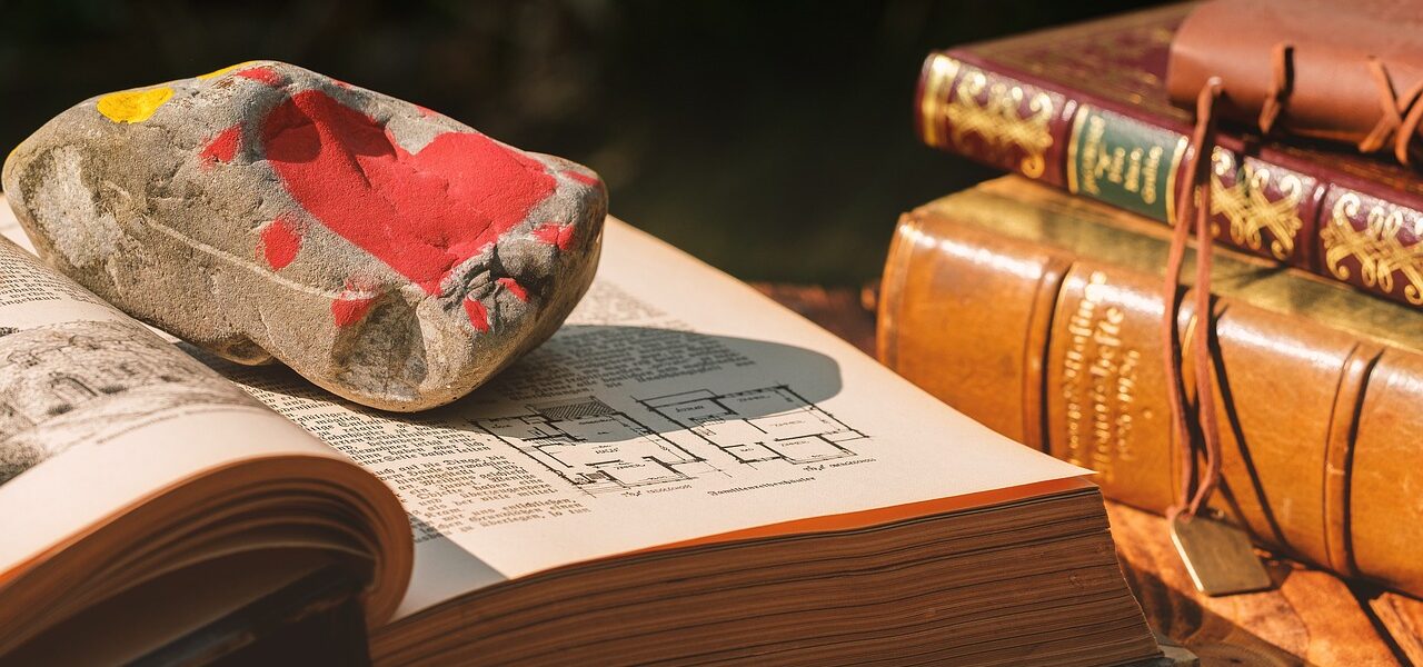 Livro aberto com uma pedra em cima pintada a superfície de vermelho. Ao lado três livros fechados, aparecendo a lombada.
