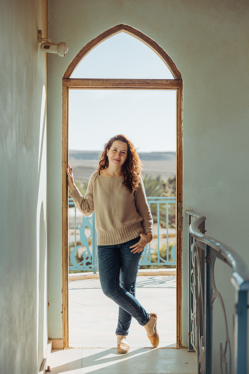 Escritora Nurit Gl de pé, na porta de uma casa em Israel com montanhas ao fundo.